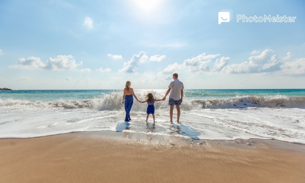 a mother and a father holding the hands of their daughter at the beach whiling walking in the water on a sunny day.