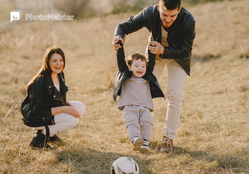 Mom kneeling in short grass. Dad holding his son up by his arms so that the son can kick a football